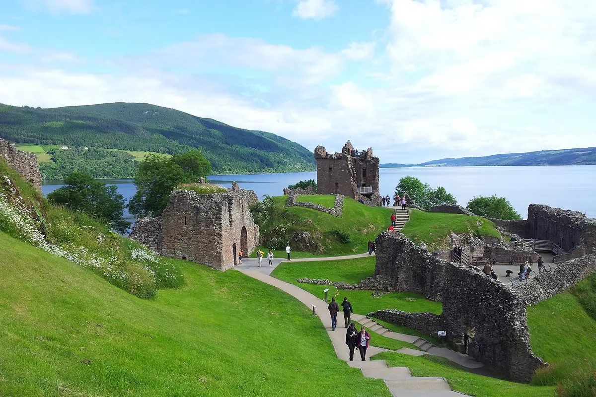 Zříceniny hradu Urquhart Castle na zeleném ostrohu u Loch Ness, s pěšinou, návštěvníky a kopci v pozadí